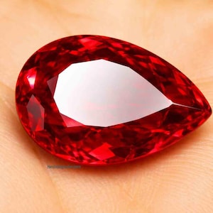Puede incluir: Una gema facetada, en forma de pera, de color rojo intenso. La gema tiene un tallado brillante con múltiples facetas que reflejan la luz. La piedra se encuentra sobre un fondo neutro, resaltando su color y forma. Es probable que la gema sea un rubí.