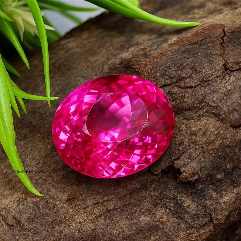 May include: A faceted, oval-shaped, hot pink gemstone. The gem is set against a dark brown, textured wooden surface, with green foliage in the background. The gemstone's cut creates multiple facets that reflect light, giving it a brilliant appearance.