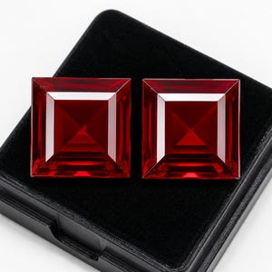 Puede incluir: Dos piedras preciosas de color rojo intenso con corte cuadrado y superficies facetadas que crean una apariencia piramidal. Las piedras están colocadas una al lado de la otra, mostrando un color carmesí translúcido y vibrante.