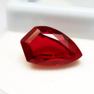 May include: A faceted, vibrant red gemstone with a unique, asymmetrical shape. The gem's polished surfaces reflect light, showcasing its clarity and rich color. The gemstone is set against a white background.