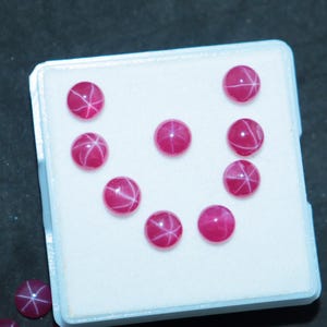 Puede incluir: Una colección de diez cabujones de rubí estrella rosa en una caja blanca. Los rubíes son redondos y tienen un patrón en forma de estrella.