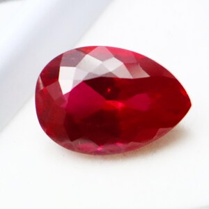 Könnte beinhalten: Ein facettierter, birnenförmiger Rubin-Edelstein. Der Edelstein hat eine tiefe, satte rote Farbe mit mehreren Facetten, die das Licht reflektieren. Der Edelstein ist vor einem weißen Hintergrund platziert.