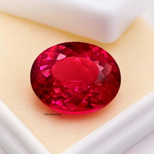 May include: A faceted, oval-shaped, deep red gemstone. The gem is set against a cream-colored background within a white box. The gem's surface reflects light, showcasing its intricate cuts. The text "Handmadejewelgems" is visible on the lower left.