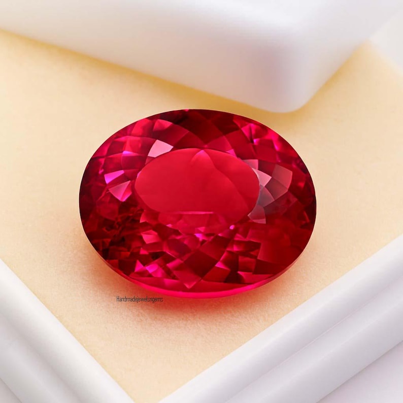 May include: A faceted, oval-shaped, deep red gemstone. The gem is set against a cream-colored background within a white box. The gem's surface reflects light, showcasing its intricate cuts. The text "Handmadejewelgems" is visible on the lower left.