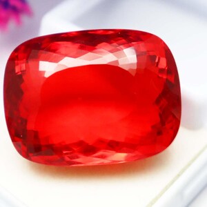 May include: A faceted, cushion-cut, red gemstone. The gem is a vibrant, transparent red with multiple facets that reflect light. The gemstone is set against a white background, highlighting its colour and clarity. This gemstone is suitable for jewellery.