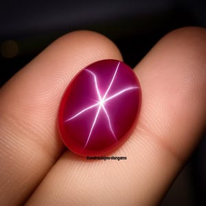 Puede incluir: Una gema de rubí ovalada y pulida con un efecto de estrella de seis rayos. La gema es de color rojo intenso con un patrón de estrella blanco brillante que emana del centro. El texto "Handmadejewelsngems" es visible en la parte inferior derecha.