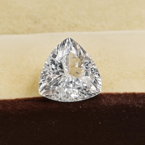 Op de afbeelding: Een heldere, driehoekig geslepen edelsteen met meerdere facetten, die het licht reflecteert. De edelsteen is geplaatst tegen een beige en bruine achtergrond, die zijn schittering toont. Het is een onderdeel van sieraden.