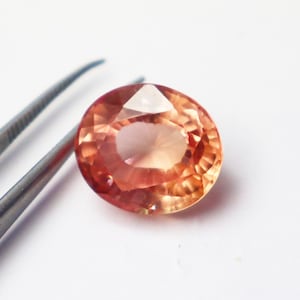 Puede incluir: Primer plano de una piedra preciosa ovalada de color melocotón con talla facetada. La piedra preciosa está sobre una superficie blanca.