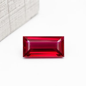 May include: A rectangular, baguette-cut gemstone with a vibrant, deep red color. The stone features precise, sharp facets that create a clear, reflective surface with internal light play, showcasing a polished finish and a symmetrical geometric shape.