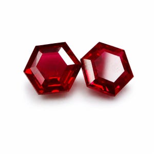 Könnte beinhalten: Zwei lose, leuchtend rote Edelsteine im Hexagonschliff. Jeder Stein weist mehrere Facetten auf, die das Licht reflektieren und ein brillantes, tiefkarmesinrotes Aussehen mit scharfen, geometrischen Kanten und einer polierten, glatten Oberfläche erzeugen.