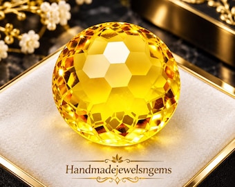May include: A faceted, spherical yellow gemstone with a honeycomb-like pattern, resting on a white velvet cushion inside a gold-trimmed box. The text "Handmadejewelsngems" is printed in gold lettering below the gemstone on the cushion.