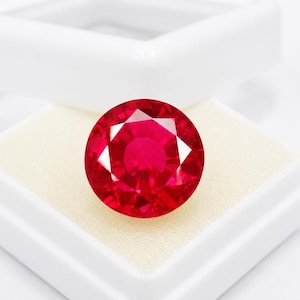 May include: A round, faceted ruby gemstone, presented in a white square box. The ruby is a deep, vibrant red colour with multiple facets that reflect light. The gemstone is set against a cream-coloured background within the box.