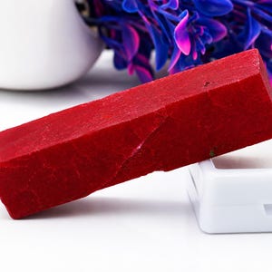 May include: A vibrant red, rectangular gemstone with a faceted surface. The gemstone is a deep, solid red color and appears to be a natural stone. It is displayed on a white surface with a white container and a blurred background.