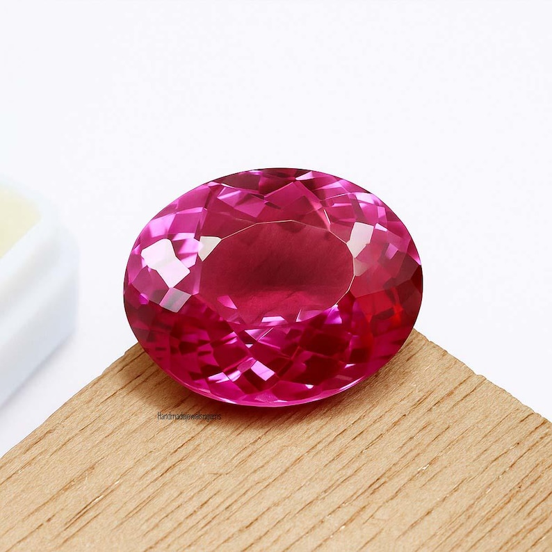 May include: A faceted, oval-cut, hot pink gemstone. The gem is set on a light brown wooden surface, with a white background. The gemstone has many facets that reflect light, creating a sparkling effect. The text "handmadejeweldesigns" is visible on the wooden surface.