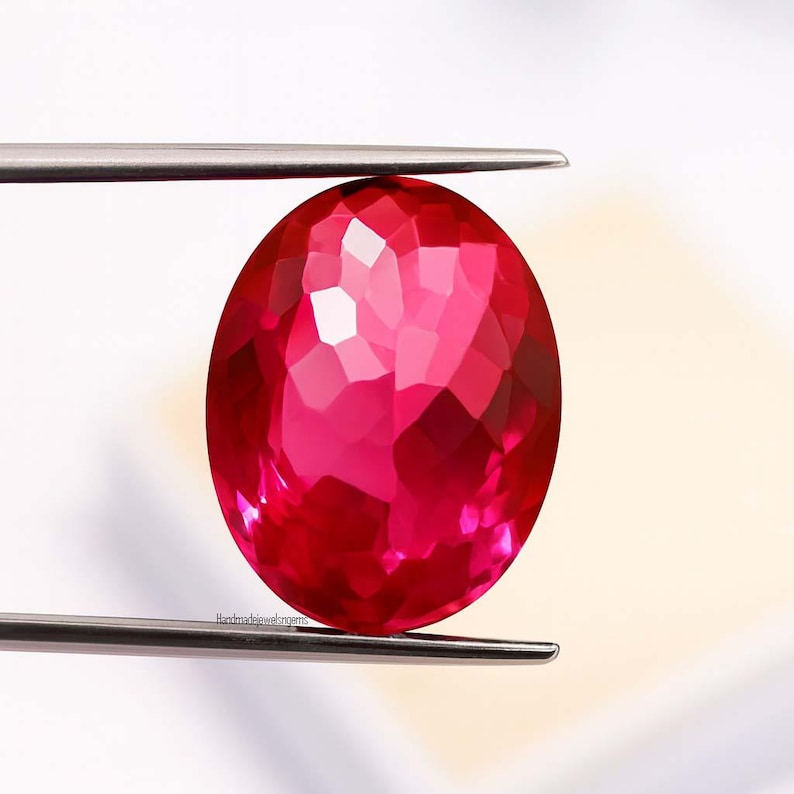 May include: A faceted, oval-shaped, ruby-red gemstone is held by silver tweezers. The gem has a brilliant cut, reflecting light across its many facets. The background is a soft, off-white. The text "handmadejewelgems" is visible on the gem.