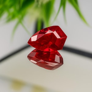 May include: A faceted, pear-shaped, deep red gemstone. The gem is highly reflective, with multiple facets catching the light. The stone is displayed on a reflective surface, creating a mirror image. The background is blurred, with green foliage.