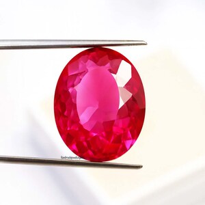 May include: A vibrant, oval-cut ruby gemstone held by tweezers. The ruby is a deep, saturated red with multiple facets that reflect light. The gemstone is set against a white background.
