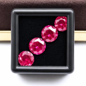 Può includere: Quattro pietre preziose rotonde, taglio brillante, di un vibrante colore rosso rosato, sono disposte in diagonale all'interno di una scatola espositiva quadrata nera. Le pietre presentano molteplici sfaccettature che riflettono la luce, creando un aspetto scintillante.