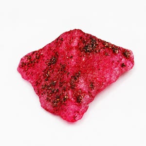 May include: A rough, irregularly shaped mineral specimen with a vibrant, translucent magenta-pink hue. The surface features a crystalline, granular texture with scattered deposits of dark, metallic-looking mineral inclusions across the top.