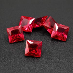May include: Six square-cut, faceted, bright red gemstones are arranged on a dark surface. The gems have sharp edges and reflect light, showcasing their clarity and vibrant colour. These could be used for jewellery making.