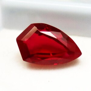 May include: A pear-shaped, faceted gemstone with a deep, vibrant red color. The stone features multiple geometric facets that reflect light, creating a brilliant, translucent appearance with sharp, defined edges and a pointed tip.