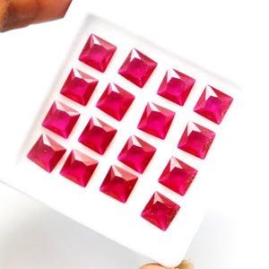 May include: A collection of 16 square-shaped ruby gemstones, arranged in rows on a white display card. The rubies are a deep red color and have a polished, faceted finish.