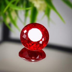 Puede incluir: Una gema roja brillante, redonda y facetada, con talla brillante. La gema es muy reflectante, con un centro blanco y un reflejo espejado debajo. El fondo está borroso, con follaje verde y una superficie gris neutra.
