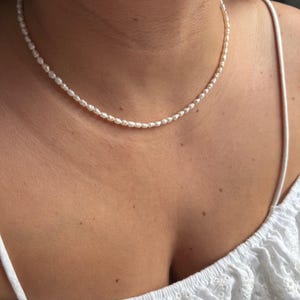 Könnte beinhalten: Zarte weiße Perlenkette. Die Halskette besteht aus kleinen, gleichmäßig verteilten Perlen, die einen klassischen und eleganten Look erzeugen. Die Perlen sind zu einem vollständigen Kreis aufgefädelt. Die Halskette wird um den Hals getragen.