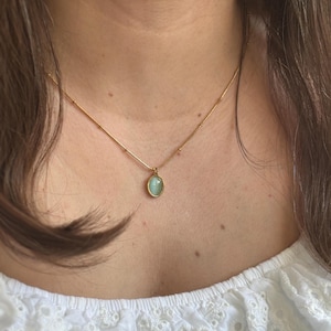 以下が含まれることがあります： 小さな楕円形のペンダントに薄い青色の宝石がついた、繊細な金のチェーンネックレスです。