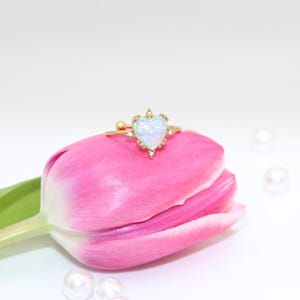 May include: A gold ring featuring a white opal heart-shaped stone surrounded by smaller stones. The ring is resting on a pink tulip.