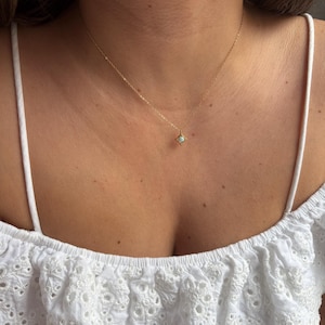 May include: A fine gold necklace with a small, square pendant holding a pale gemstone. The necklace is set against skin and a white, broderie anglaise top. The pendant is the central feature of this elegant piece of jewellery.