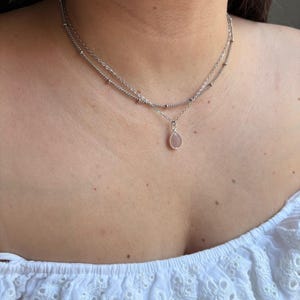 May include: A double-layered silver-toned necklace featuring a delicate chain with small metallic beads and a second chain with a single teardrop-shaped pendant containing a pale pink stone, resting against a white eyelet fabric garment.