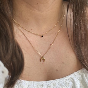 Può includere: Due collane d'oro sovrapposte sul collo di una persona. La collana superiore ha un piccolo ciondolo quadrato nero. La collana inferiore ha un ciondolo a forma di mezzaluna.