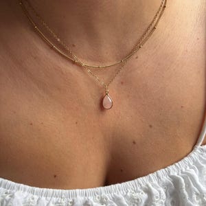Puede incluir: Un delicado collar dorado con dos cadenas. La cadena superior tiene pequeñas cuentas, y la cadena inferior presenta un colgante de piedra preciosa rosa pálido en forma de lágrima. El collar se lleva sobre una piel clara y una blusa blanca.