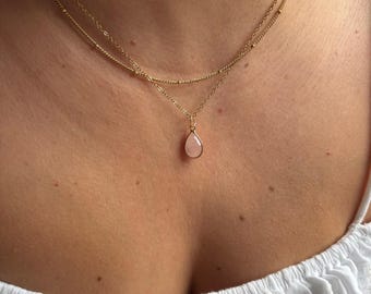 Könnte beinhalten: Eine mehrlagige Goldhalskette mit zwei zarten Ketten. Die obere Kette ist schlicht gehalten, während die untere Kette einen einzelnen, kleinen, blassrosa tropfenförmigen Edelsteinanhänger in einer dünnen Goldfassung trägt.