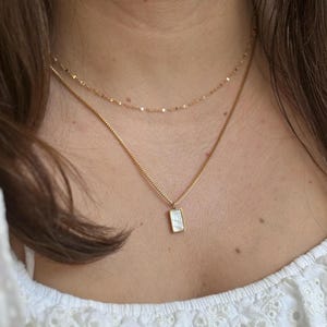 May include: A gold chain necklace with a small rectangular pendant featuring a white iridescent stone. The necklace is layered over a thinner gold chain.