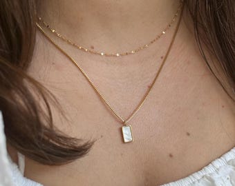 May include: A gold chain necklace with a small rectangular pendant featuring a white iridescent stone. The necklace is layered over a thinner gold chain.