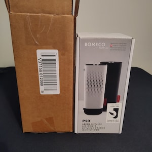 Könnte beinhalten: Ein weißer und schwarzer Aromadiffusor und Luftbefeuchter mit der Marke "BONECO" und der Modellnummer "P50". Das Produkt wird mit einem USB-C-Kabel betrieben. Die Box ist mit "healthy air" und "TRADITION SINCE 1956" beschriftet.
