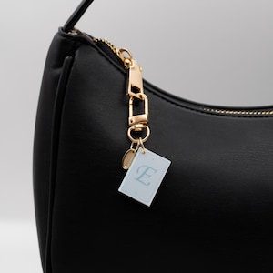 May include: Black shoulder bag with a gold zip and chain. A small tag with the letter "E" hangs from the zip. The bag has a curved shape and a strap.