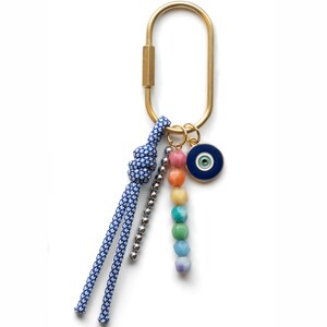 Könnte beinhalten: Goldfarbener Metall-Karabiner-Schlüsselanhänger mit blau-weiß gemustertem Seil, einer Kette aus silbernen Perlen und einer Regenbogenfarbenen Perlenkette. Ein blauer und goldener Evil-Eye-Anhänger ist ebenfalls angebracht.