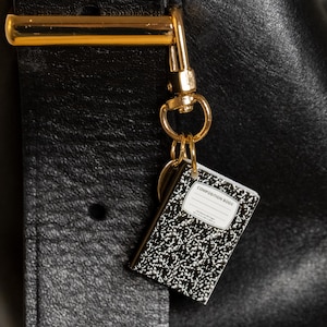Puede incluir: Un llavero de cuaderno de composición en miniatura con una cubierta moteada en blanco y negro, colgando de un cierre dorado. El llavero está sujeto a un bolso de cuero negro.