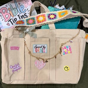 May include: A beige canvas tote bag featuring a colorful crochet strap, a gold chain with charms, and several patches including text that reads Saved by Grace, Faith, Created with a purpose, and Love like Jesus, along with a yellow smiley face patch.