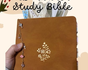 Könnte beinhalten: Eine hellbraune ESV Loaded Studienbibel mit einem goldfarbenen Blumenmotiv auf dem Einband. Das Buch ist mit einer goldfarbenen Kette mit verschiedenen kleinen Anhängern an der Seite sowie mehreren bunten Seitenreitern und Bändern versehen.