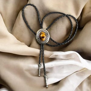 May include: A bolo tie with a silver-toned circular pendant featuring an oval, amber-colored stone. The pendant is surrounded by an ornate design. The black braided cord has decorative silver-toned tips. The tie is displayed on a beige and white fabric.