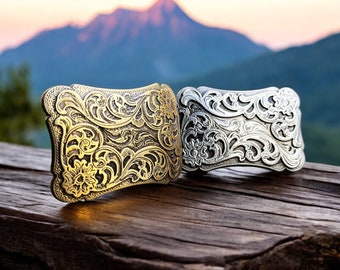 May include: Two ornate metal belt buckles, one gold and one silver, with intricate floral designs. The buckles are on a wooden surface.