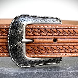 May include: Close-up of a brown leather belt with a decorative silver buckle. The belt features a detailed woven pattern and white stitching. The buckle has an ornate floral design.