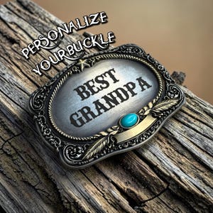 Może przedstawiać: Prostokątna klamra do paska w kolorze srebrnym z ozdobną krawędzią, zawierająca napis "BEST GRANDPA" czarnymi literami, małą gwiazdkę na górze, pojedynczy owalny kamień w kolorze turkusowym oraz grawerunki przypominające pióra. Widoczny jest tekst "PERSONALIZE YOUR BUCKLE".