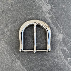 May include: A silver metal belt buckle with a rounded, horseshoe-shaped design.