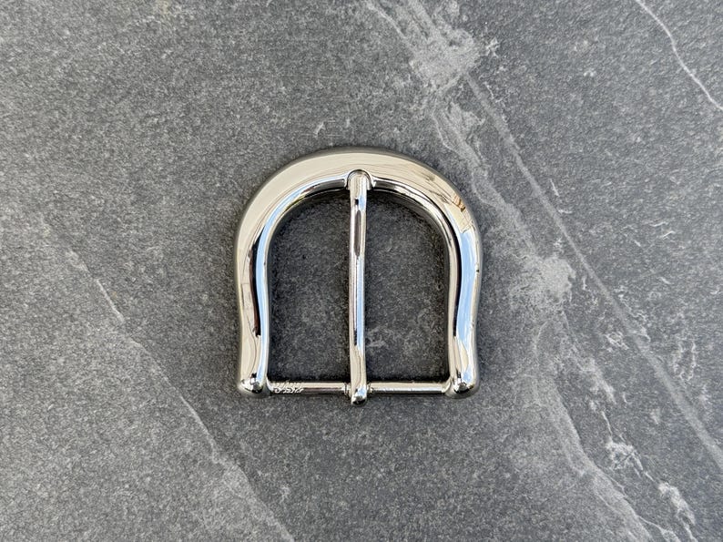 May include: A silver metal belt buckle with a rounded, horseshoe-shaped design.