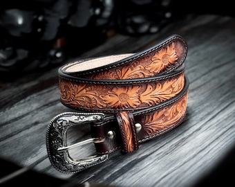 May include: A brown leather belt with a silver buckle. The belt is decorated with an intricate floral pattern.